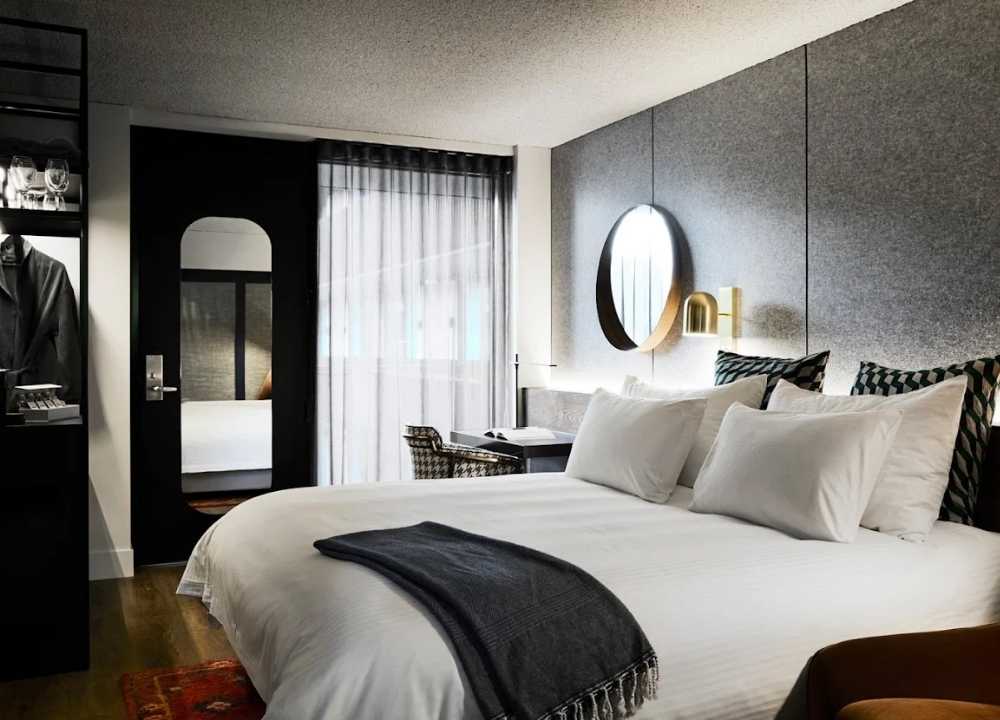 A stylish and contemporary guest room at Zagame's House in Melbourne, featuring a plush king-size bed with crisp white linens, patterned cushions, and a dark throw blanket. The room is decorated with modern furnishings, including a circular mirror with a backlight, a sleek desk and chair, and minimalist lighting. Floor-to-ceiling curtains filter natural light, creating a warm and inviting atmosphere.